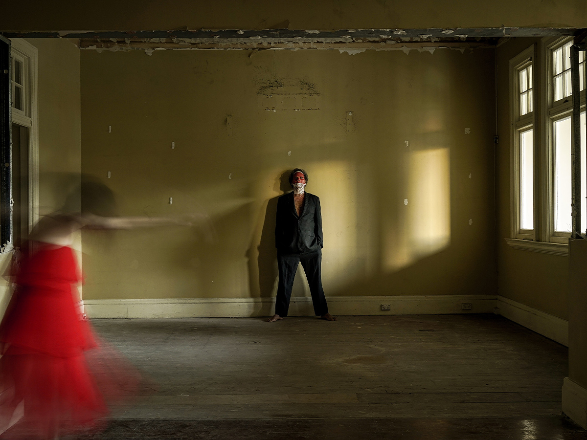 In a dimly lit, sparse room with peeling paint, an Indigenous man stands barefoot and poised against the wall, wearing a suit and traditional face paint. A figure in a vibrant red dress moves dynamically in the foreground, creating a blur of motion. Sunlight streams through the windows, casting dramatic shadows. This evocative scene is part of the *All Come Under* exhibition at the Court House Gallery, highlighting the collaboration and creative expression of artists Darren Blackman, Zane Saunders, and Bonemap.